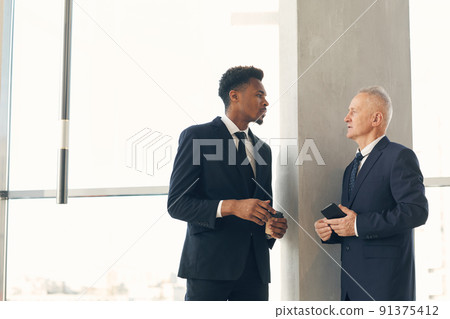 Serious thoughtful multi-ethnic businessmen in dark formal suits standing in lobby and brainstorming about new strategy for project implementation Serious thoughtful multi-ethnic businessmen in dark formal suits standing in lobby and brainstorming about new strategy for project implementation 91375412