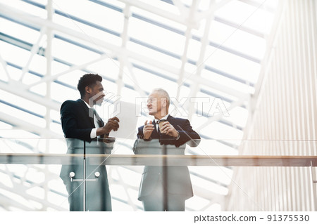 Young Afro-American specialist with papers standing in hall and interacting with senior business executive on contract issues 91375530