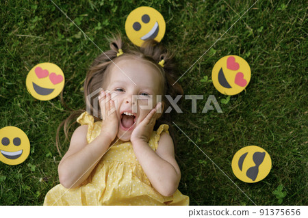 A funny little girl surrounded by a variety of paper emoticons looks up in surprise with her mouth open in delight. Emotional little kid celebrates world smile day 91375656