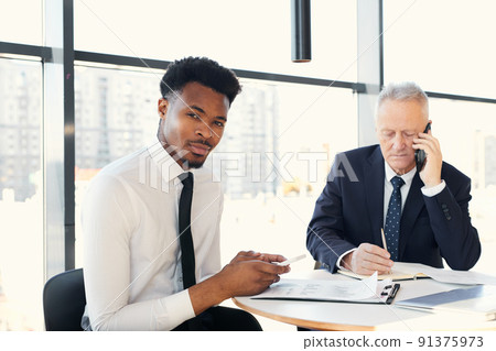Portrait of content confident young black businessman with beard using smartphone for notes while working on papers with mature colleague 91375973