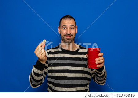 Smiling latin man with a beard dressed in a striped sweater putting a euro coin in a metal piggy bank, trying to save so he can travel on vacation. Isolated on blue studio background. Smiling latin man with a beard dressed in a striped sweater putting a euro coin in a metal piggy bank, trying to save so he can travel on vacation. Isolated on blue studio background. 91376819