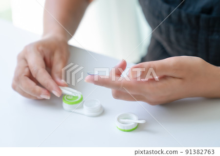 Woman puts it down contact lens on her index finger with tweezers. There is container for lenses nearby. Close-up, side view Woman puts it down contact lens on her index finger with tweezers. There is container for lenses nearby. Close-up, side view 91382763
