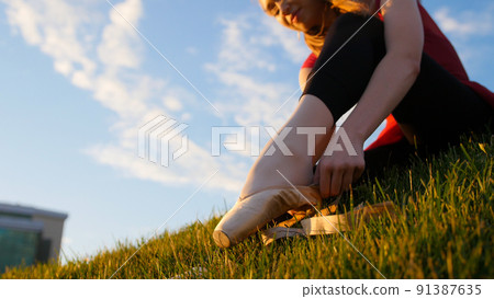 Young woman put on her pointe shoes - sitting on the green grass at sunset Young woman put on her pointe shoes - sitting on the green grass at sunset 91387635