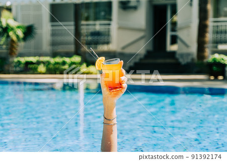 Closeup woman's hand sticking out of swimming pool holding tropical cocktail. Cropped girl's female's hand with an orange lemonade and the hotel pool in the background. Hello summer holiday vacation. 91392174