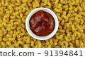 Saucer with delicious red ketchup on background of many dry pasta in form of spirals. Selective focus. Food background. Top view. 91394841