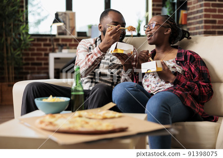 Relaxed partners eating noodles from takeout delivery box, having fun with fast food takeaway meal on couch. Using chopsticks to eat dinner and watching movie or film on television program. Relaxed partners eating noodles from takeout delivery box, having fun with fast food takeaway meal on couch. Using chopsticks to eat dinner and watching movie or film on television program. 91398075
