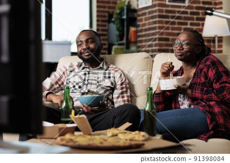 African american couple eating takeaway meal with noodles and chips to watch movie on television. Leisure activity together with film on tv and fast food delivery package, having fun. African american couple eating takeaway meal with noodles and chips to watch movie on television. Leisure activity together with film on tv and fast food delivery package, having fun. 91398084