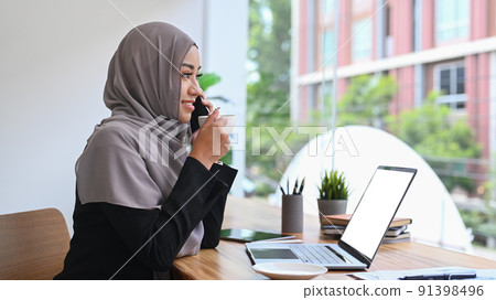Muslim woman wearing headscarf talking on phone and drinking hot coffee in modern office room 91398496