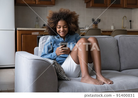 Cute African teenage girl sit on sofa use smart phone 91400831