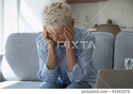 Upset older woman holding head with hands seated on sofa 91401078