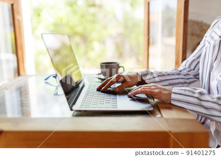 Cropped shot of young businesswoman working on her laptop while sitting at a cafe place or home, next to window. Cropped shot of young businesswoman working on her laptop while sitting at a cafe place or home, next to window. 91401275