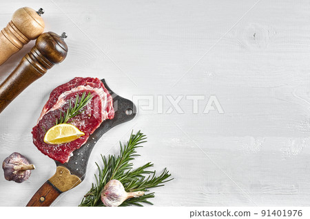 Fresh raw steaks with herbs, garlic, olive oil, pepper, salt, rosemary and a kitchen ax on white background. Top view. 91401976
