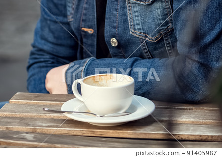 Person in jean jacket sits in outdoor cafe with empty cup of coffee on table top 91405987