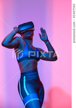 Modern african black woman with short hiar in sport top and leggings wearing glasses of virtual reality in blue and pink neon light Modern african black woman with short hiar in sport top and leggings wearing glasses of virtual reality in blue and pink neon light 91407565