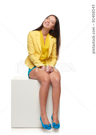 Full length of smiling girl in yellow and blue clothes sitting on white cube dreaming with closed eyes Full length of smiling girl in yellow and blue clothes sitting on white cube dreaming with closed eyes 91408545