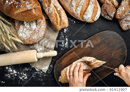 Woman hands cutting a loaf of bread on rustic wooden board, with wheat ears and knife, top view. 91411907