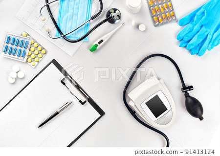 White electric tonometer with stethoscope, multicolored pills, mask and thermometer on white table. Top view 91413124