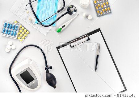 White electric tonometer with stethoscope, multicolored pills, mask and thermometer on white table. Top view 91413143