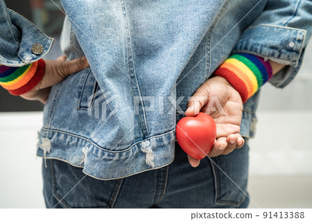 Asian lady wearing rainbow flag wristbands and hold red heart, symbol of LGBT pride month celebrate annual in June social of gay, lesbian, bisexual, transgender, human rights. Asian lady wearing rainbow flag wristbands and hold red heart, symbol of LGBT pride month celebrate annual in June social of gay, lesbian, bisexual, transgender, human rights. 91413388