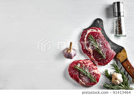 Two fresh raw steaks with herbs, garlic, olive oil, pepper, salt, rosemary and a kitchen ax on white background. Top view. 91413464