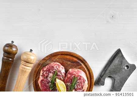 Two raw beef steak with spices on wooden board and kitchen ax on white background, top view 91413501