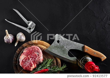 Piece of beef tenderloin, with ax for cutting and chopping meat, spices were cooking - rosemary, pepper, salt, garlic. On old wooden board on stone black table. 91413577