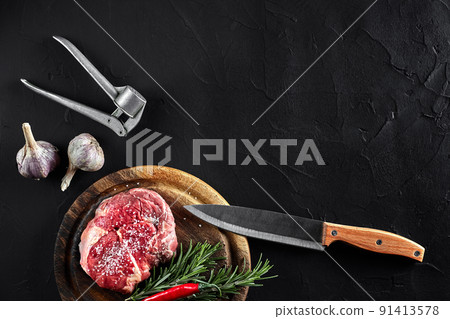Piece of beef tenderloin, with knife for cutting and chopping meat, spices were cooking - rosemary, pepper, salt, garlic. On old wooden board on stone black table. 91413578