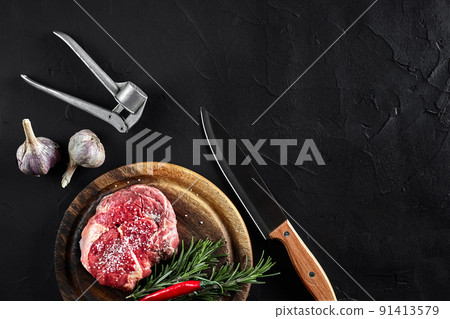 Piece of beef tenderloin, with knife for cutting and chopping meat, spices were cooking - rosemary, pepper, salt, garlic. On old wooden board on stone black table. 91413579