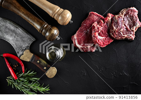 Piece of beef tenderloin, with ax for cutting and chopping meat, spices were cooking - rosemary, pepper on old black table. Top view copy space Piece of beef tenderloin, with ax for cutting and chopping meat, spices were cooking - rosemary, pepper on old black table. Top view copy space 91413856