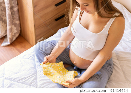 Pregnancy baby clothes. Beautiful pregnant mother with yellow baby clothes. Pregnancy woman hugging belly and packing maternity hospital bag. Concept maternity, pregnancy, childbirth. Pregnancy baby clothes. Beautiful pregnant mother with yellow baby clothes. Pregnancy woman hugging belly and packing maternity hospital bag. Concept maternity, pregnancy, childbirth. 91414424