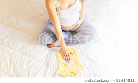 Woman baby clothes. Happy young pregnancy mother holding yellow baby clothes. Pregnant woman packing suitcase, bag for maternity hospital. Concept of pregnancy, maternity, expectation for baby birth. 91414811