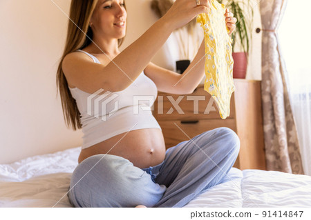 Woman baby clothes. Happy young pregnancy mother holding yellow baby clothes. Pregnant woman packing suitcase, bag for maternity hospital. Concept of pregnancy, maternity, expectation for baby birth. 91414847