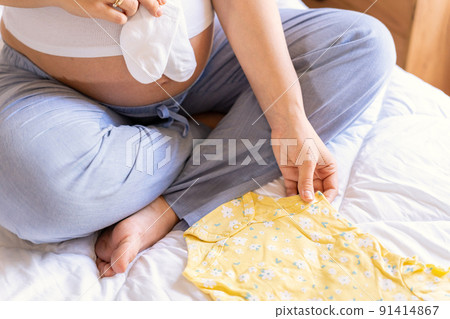 Woman baby clothes. Happy young pregnancy mother holding yellow baby clothes. Pregnant woman packing suitcase, bag for maternity hospital. Concept of pregnancy, maternity, expectation for baby birth. Woman baby clothes. Happy young pregnancy mother holding yellow baby clothes. Pregnant woman packing suitcase, bag for maternity hospital. Concept of pregnancy, maternity, expectation for baby birth. 91414867