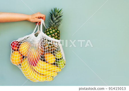 Top view female hand holding string bag, mesh bag, grocery bag with organic fresh exotic fruits on light green background. Zero waste, eco-friendly shopping. Healthy diet. Flat Lay Top view female hand holding string bag, mesh bag, grocery bag with organic fresh exotic fruits on light green background. Zero waste, eco-friendly shopping. Healthy diet. Flat Lay 91415013