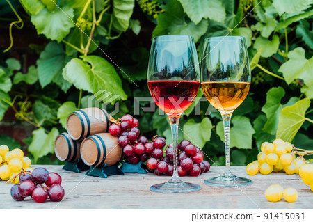 Winery concept. Glasses of white and red wine with miniature wine barrels on the wooden table with grape berries on background of vineyards. Wine tasting, Degustation. Selective focus, copy space. Winery concept. Glasses of white and red wine with miniature wine barrels on the wooden table with grape berries on background of vineyards. Wine tasting, Degustation. Selective focus, copy space. 91415031