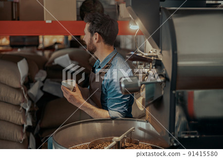 Business owner with cup of fresh coffee on background of roasting machine looking away 91415580