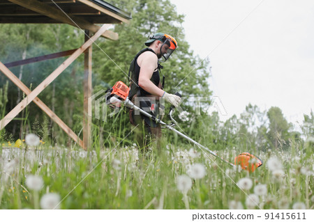 Man mowing tall grass with petrol lawn trimmer in the garden or backyard. Process of lawn trimming with hand mower Man mowing tall grass with petrol lawn trimmer in the garden or backyard. Process of lawn trimming with hand mower 91415611