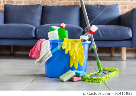 Bucket with bottles of detergent, cleaning tools and mopping stick in front of sofa in living room. Housework cleaning equipment. Bucket with bottles of detergent, cleaning tools and mopping stick in front of sofa in living room. Housework cleaning equipment. 91415922