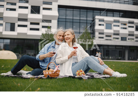 Caucasian elderly couple sitting on a blanket on a grass in summer. Man and woman drinking a wine. Woman wearing white shirt and man blue one. 91418285