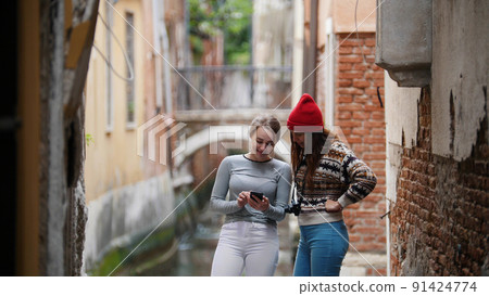 Two young women standing near the water channel and looking at the phone screen - Venice, Italy 91424774