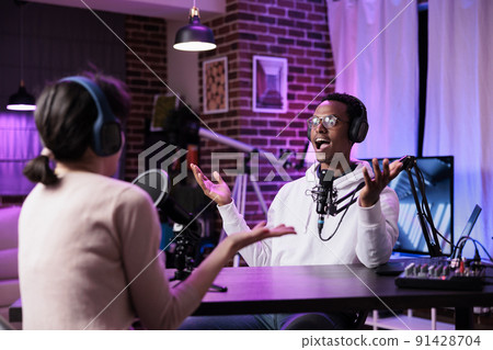 Podcast presenter recording livestream chat at production station, using modern equipment for audience. Male influencer interviewing woman for online broadcast discussion in studio. 91428704