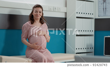 Portrait of future mother with baby bump sitting on cabinet bed, waiting to receive health care consultation from medic. Pregnant woman attending checkup appointment in office. Portrait of future mother with baby bump sitting on cabinet bed, waiting to receive health care consultation from medic. Pregnant woman attending checkup appointment in office. 91428743