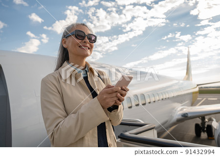 Cheerful woman with phone standing outdoors at airport and looking away 91432994