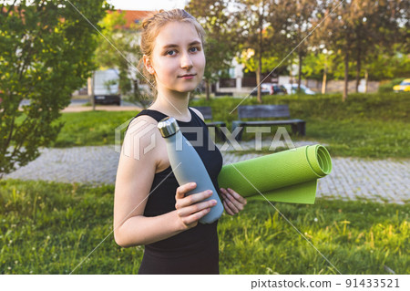 Happy young blonde woman holds yoga mat and water bottle in park. Healthy lifestyle, sport, fitness Happy young blonde woman holds yoga mat and water bottle in park. Healthy lifestyle, sport, fitness 91433521
