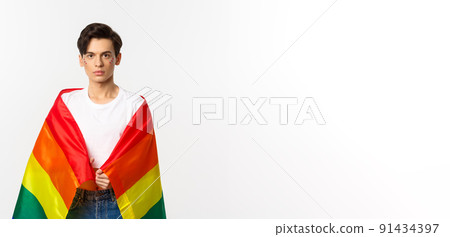 Image of serious-looking gender fluid person with glitter on face, wearing lgbtq rainbow flag, standing over white background 91434397