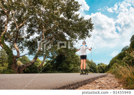A sporty man in a helmet trains on roller skis on a country road, rear view. Biathlon race with ski poles. Low angle view. Blue sky and beautiful nature along the road. The concept of summer workout A sporty man in a helmet trains on roller skis on a country road, rear view. Biathlon race with ski poles. Low angle view. Blue sky and beautiful nature along the road. The concept of summer workout 91435949