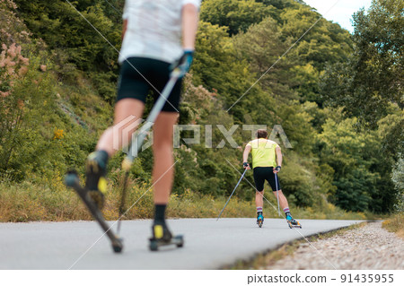 Biathlon workout. Two athletic men training on the roller ski at country road, back view. Low angle view. Concept of sports competition and summer activity 91435955