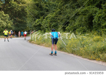 Roller ski olympiad. Group of athletes training before the competition. View from the back. Biathlon ride on the roller skis with ski poles, in the helmet. Concept of sport and summer contest 91435959