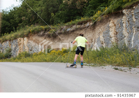 Hard training an athlete on the roller ski, back view. Biathlon ride on the roller skis with ski poles, in the helmet. Summer workout 91435967