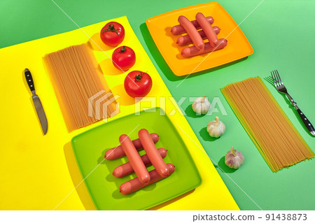 A lot of raw sausages on plate. On green and yellow background with pasta and vegetables, top view. 91438873